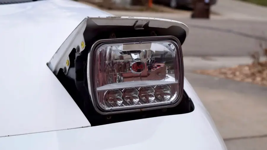 A close-up of a car's pop-up headlight, partially raised, showing a modern LED light unit installed in an older vehicle. The car is white and parked outdoors, with a blurred background.