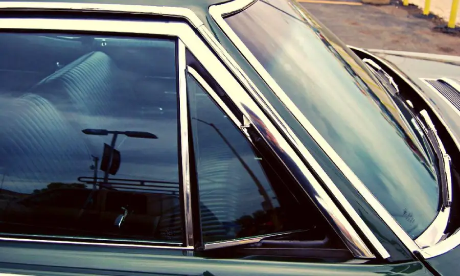 Close-up of a classic car’s side window with chrome trim, green paint, and striped upholstery visible inside. The reflection shows a streetlamp and parking lot in the background.