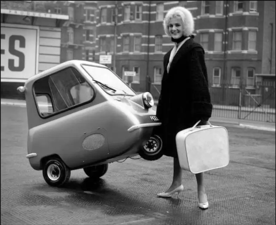 A woman in a coat and heels holds a suitcase and lifts a tiny three-wheeled car by its rear, smiling, on a city street with brick buildings in the background.