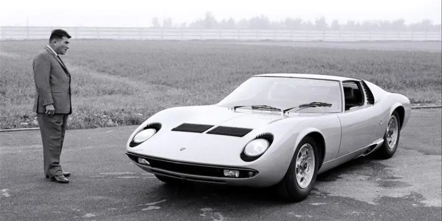 A man in a suit stands on a paved area, looking at a classic Lamborghini Miura parked in front of him, with grass and a fence in the background.