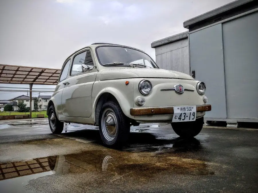 A classic white Fiat 500 is parked on wet pavement, reflecting in a small puddle. A modern building and a metal carport are in the background under a cloudy sky.