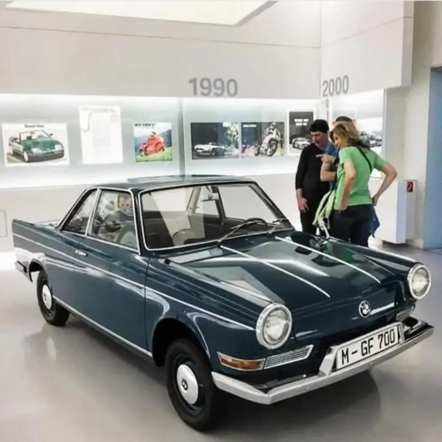 A vintage blue coupe car is displayed indoors in front of a wall with automotive photos and the years 1990 and 2000. Three people stand to the right, looking at the car.