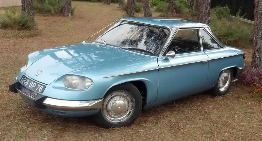 A light blue vintage coupe car is parked on grass among pine trees, with chrome details and a distinctively rounded front design.