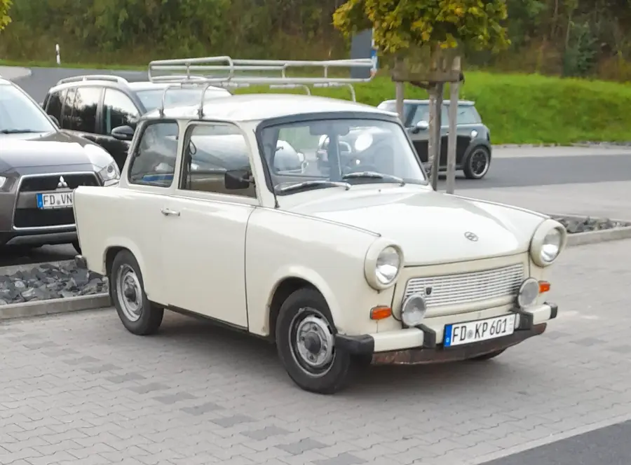 A white vintage Trabant car with a roof rack is parked on a paved area near other cars, beside a small tree, in an outdoor setting.