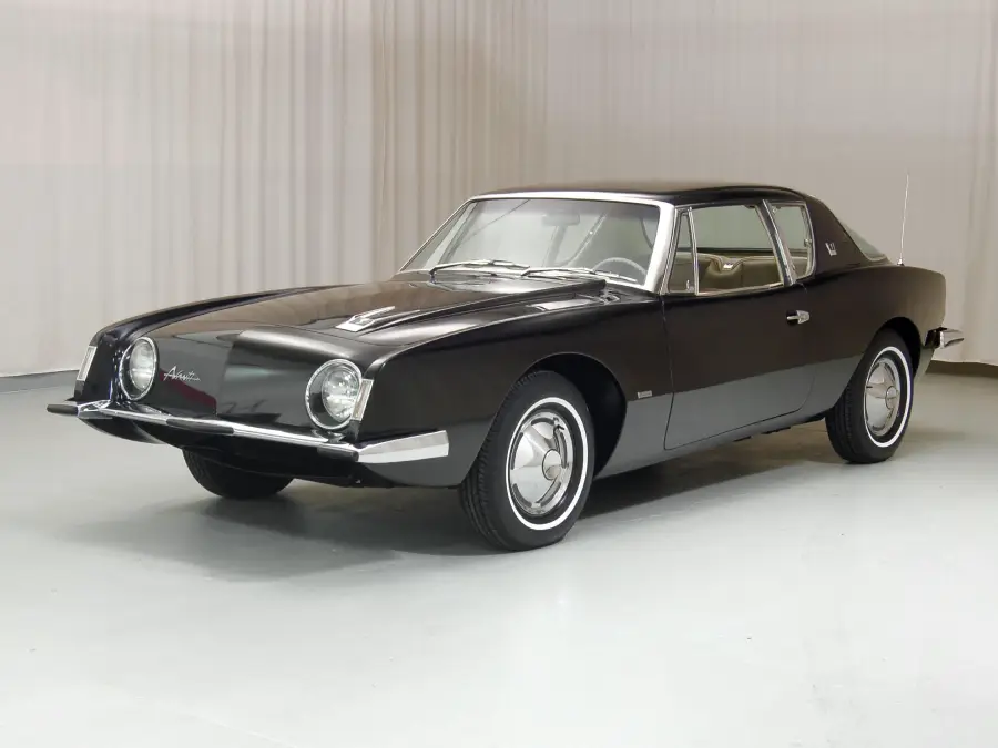 A vintage black Studebaker Avanti car with whitewall tires is parked indoors on a light-colored floor, with plain white curtains in the background.