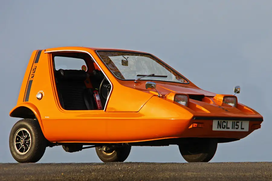 An orange three-wheeled car with two front wheels, one rear wheel, and a single side door is parked on a paved surface against a plain background. The license plate reads "NGL 115 L.