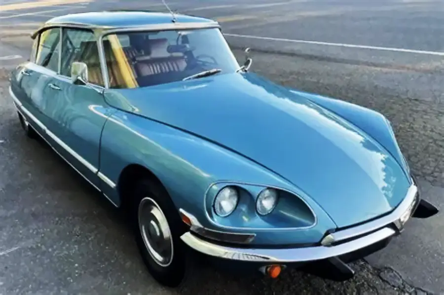 A vintage blue Citroën DS car is parked on a street, showing its unique aerodynamic shape, round headlights, and chrome detailing.