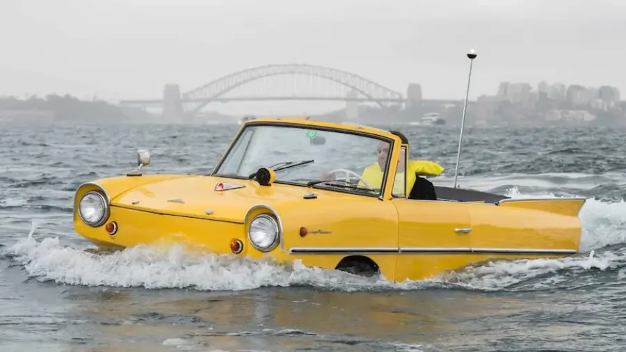 A yellow amphibious car drives on water with a person inside, city buildings and a large bridge are visible in the foggy background.