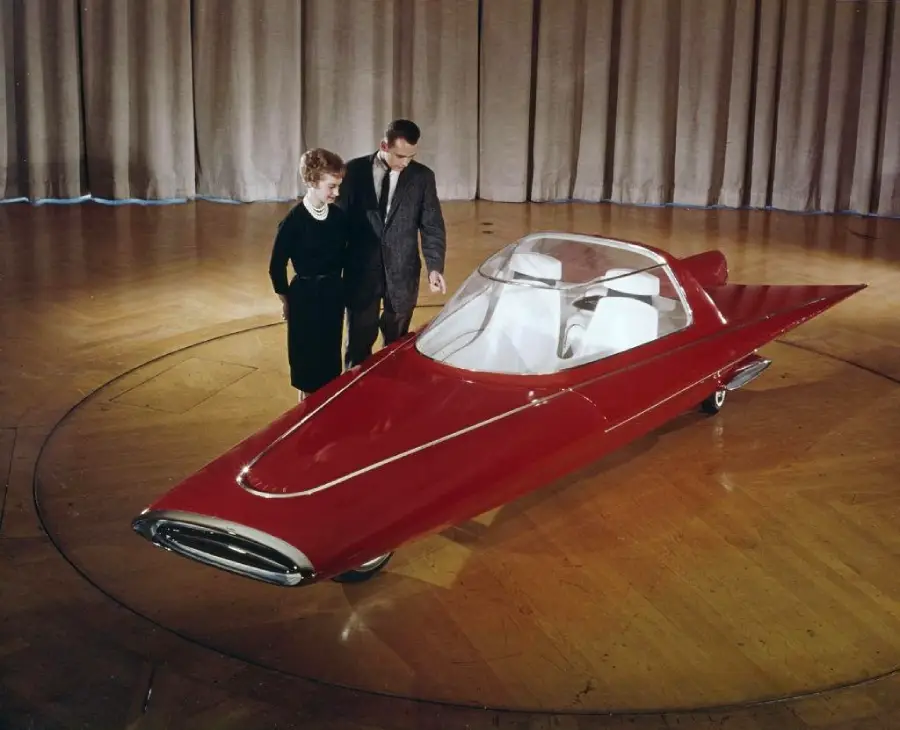 A woman and a man stand beside a futuristic, red, single-seat concept car with a clear canopy, displayed indoors on a wood floor with closed beige curtains in the background.