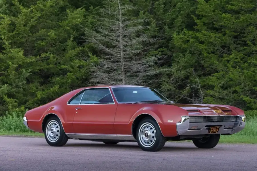 A vintage red coupe car with shiny chrome details is parked on a paved road. Lush green trees fill the background.