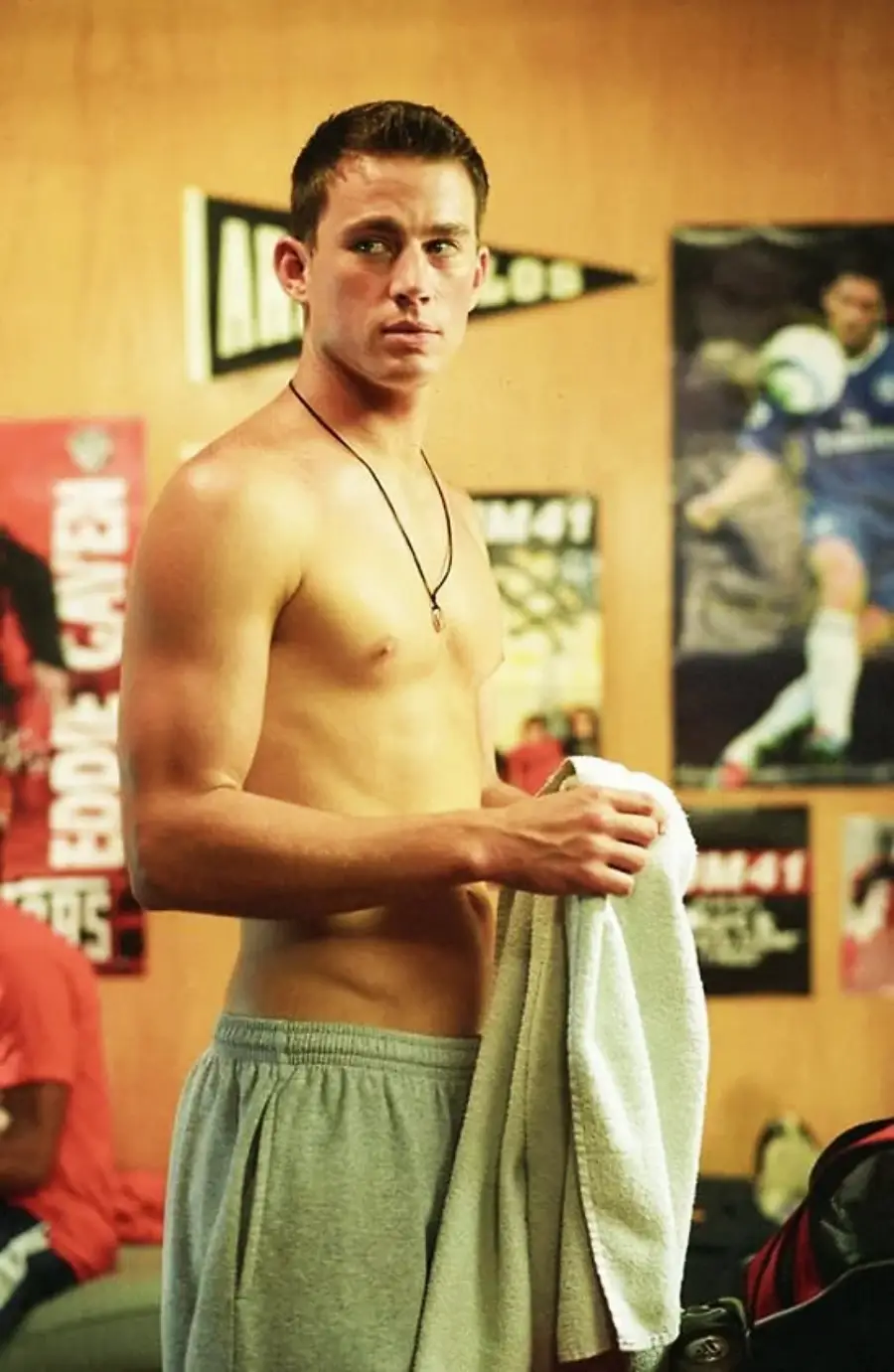 A young man stands shirtless in a locker room, holding a towel. He wears gray shorts and a necklace. Sports posters decorate the wooden wall behind him.