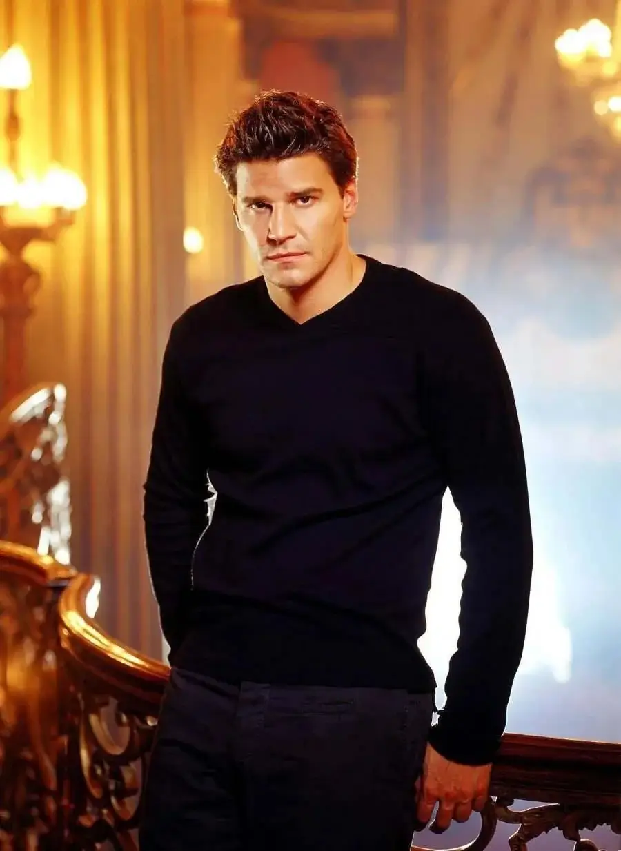 A man with short dark hair, wearing a black long-sleeve shirt, stands by an ornate staircase in a warmly lit, elegant room with golden tones and soft lighting.