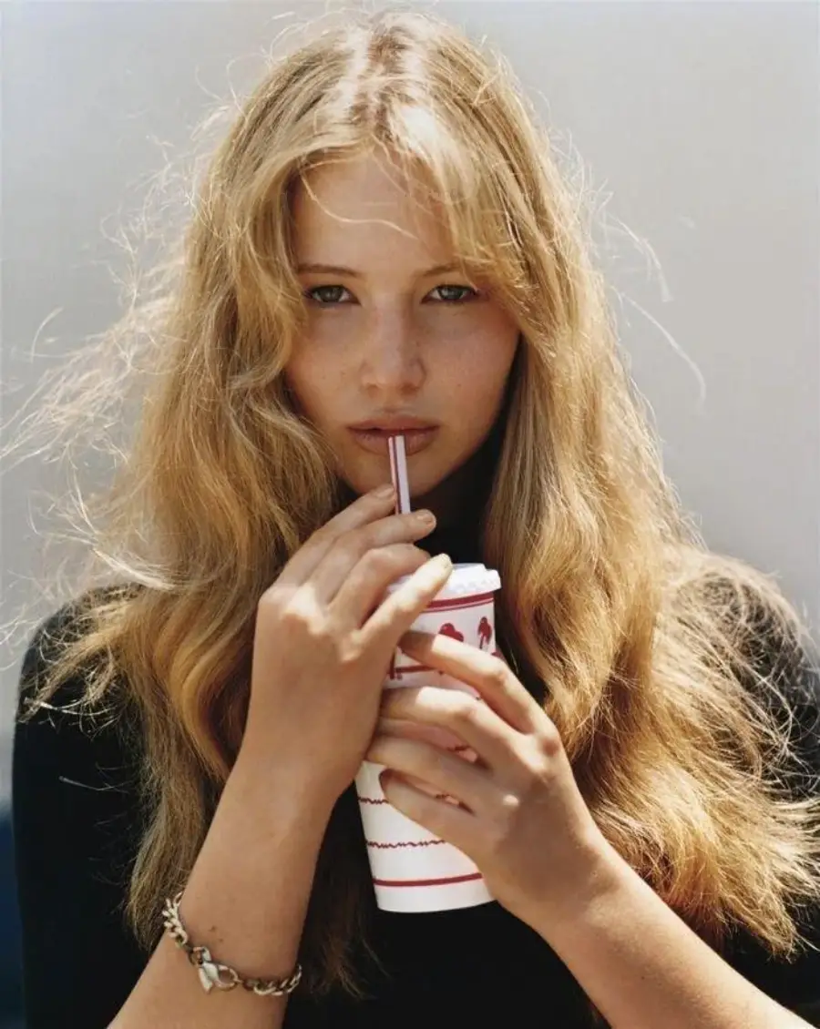 A young woman with long, wavy blonde hair and a black top drinks from a cup with red stripes, looking at the camera, against a plain background.