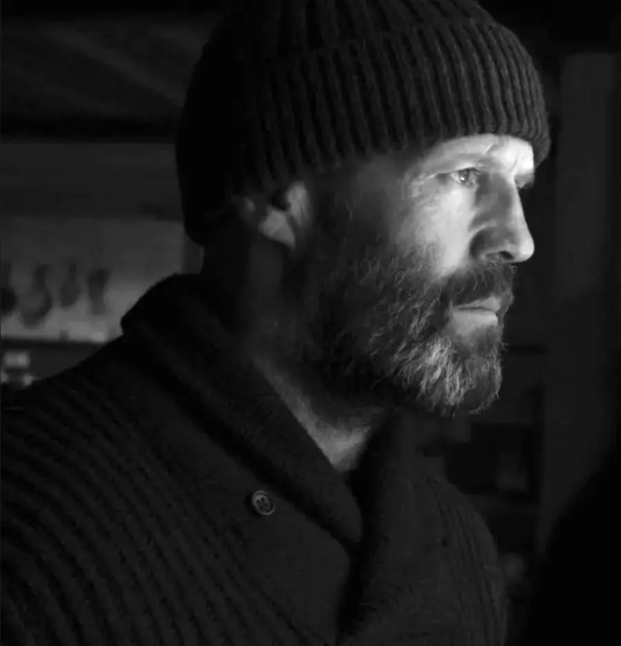 A bearded man in a knit cap and sweater looks thoughtfully to the side in a black and white photo, with dramatic lighting highlighting his profile.