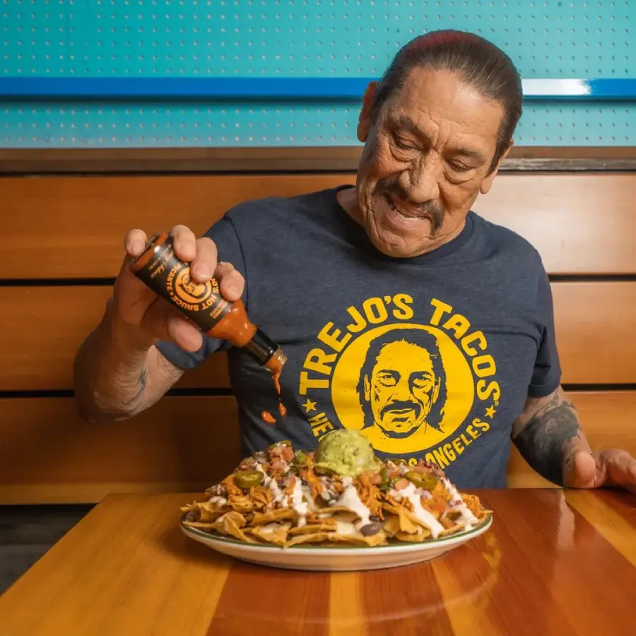 A man in a "Trejo's Tacos" shirt pours hot sauce onto a large plate of loaded nachos with guacamole, sitting at a wooden table in a restaurant.