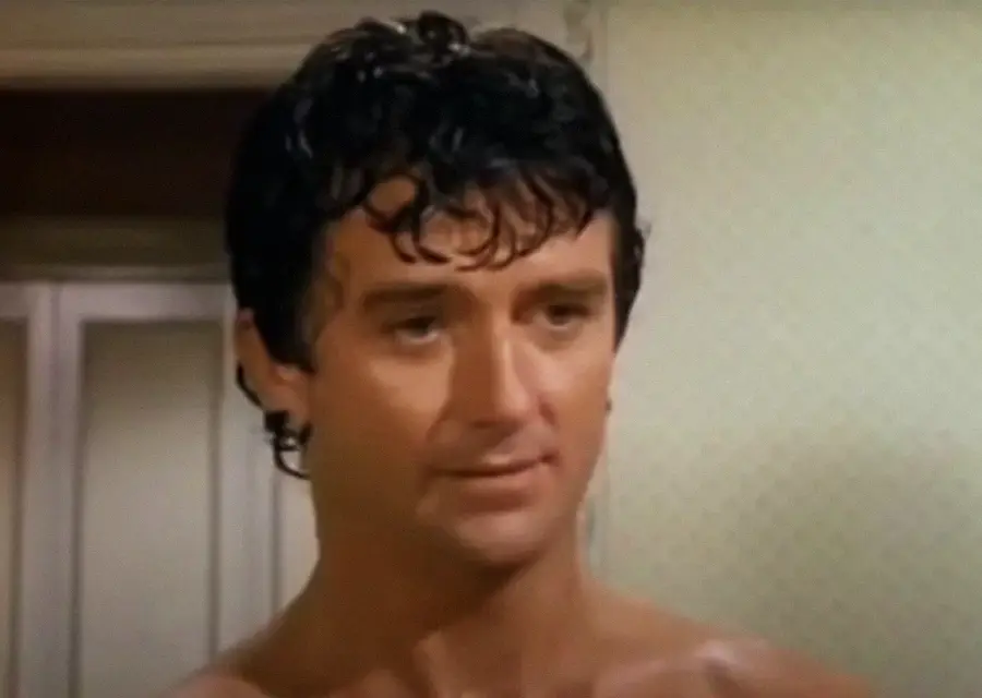 A man with short, curly dark hair stands indoors, looking slightly to the side with a neutral expression. The background shows a light-colored wall and a doorway.