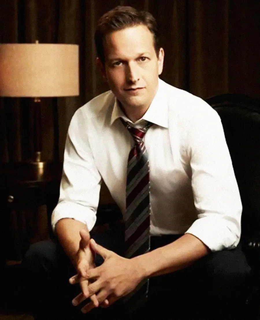 A man in a white shirt and striped tie sits on a chair, hands clasped, in a dimly lit room with a lamp in the background.