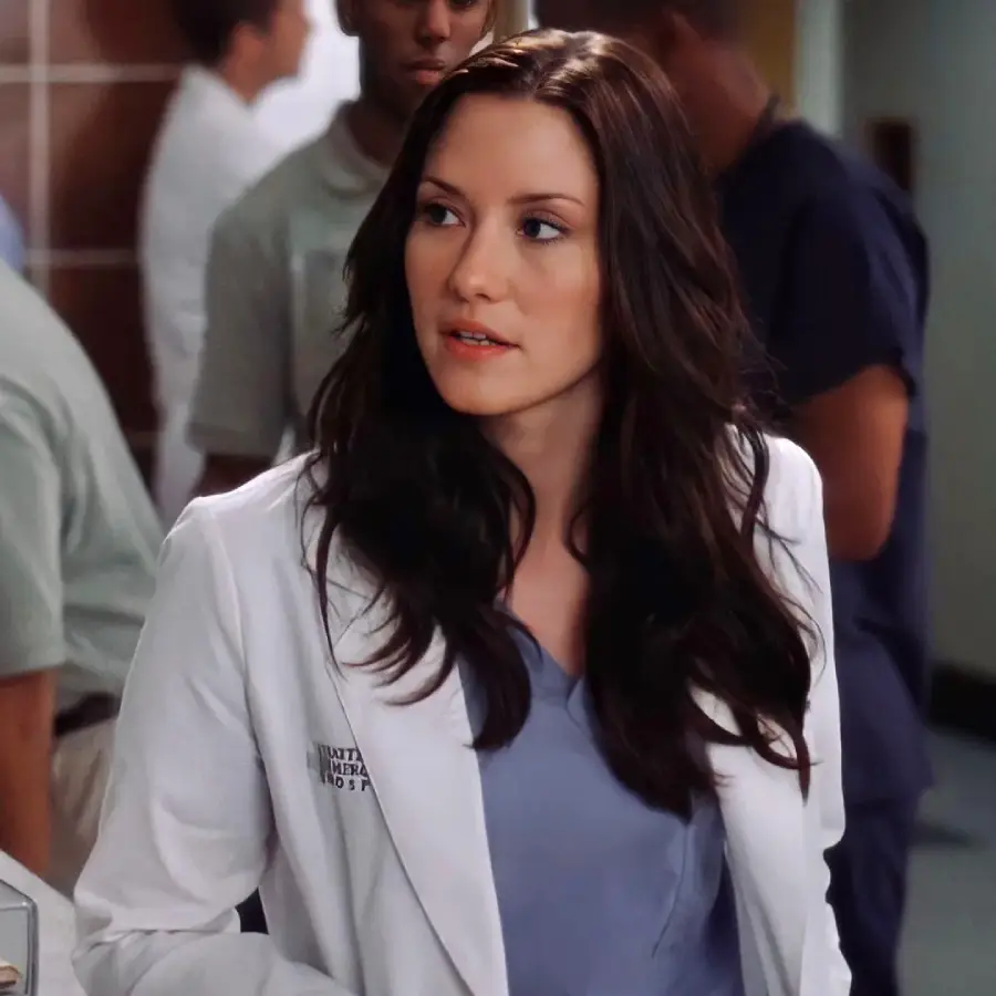 A woman with long dark hair in a white lab coat stands indoors, looking to the side. Other people in medical scrubs are in the blurred background, suggesting a hospital setting.