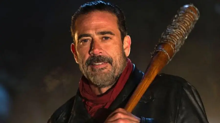 A man with a beard and slicked-back hair holds a barbed wire-wrapped baseball bat over his shoulder, wearing a red scarf and black leather jacket, standing in dim lighting.