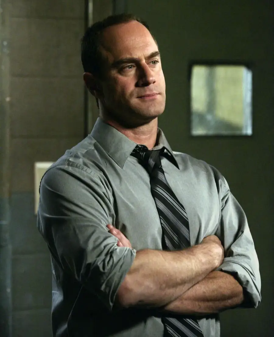 A man in a gray dress shirt and striped tie stands with his arms crossed, looking thoughtful. He is in a dimly lit room with a small window in the background.