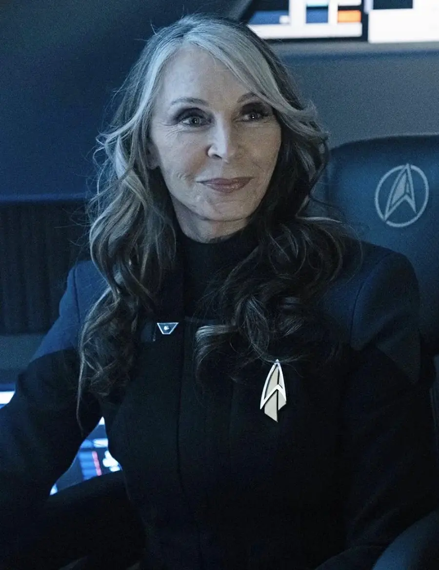 A woman with long wavy hair, wearing a black uniform with a silver Star Trek insignia, sits in a futuristic chair on a spaceship bridge, smiling softly.
