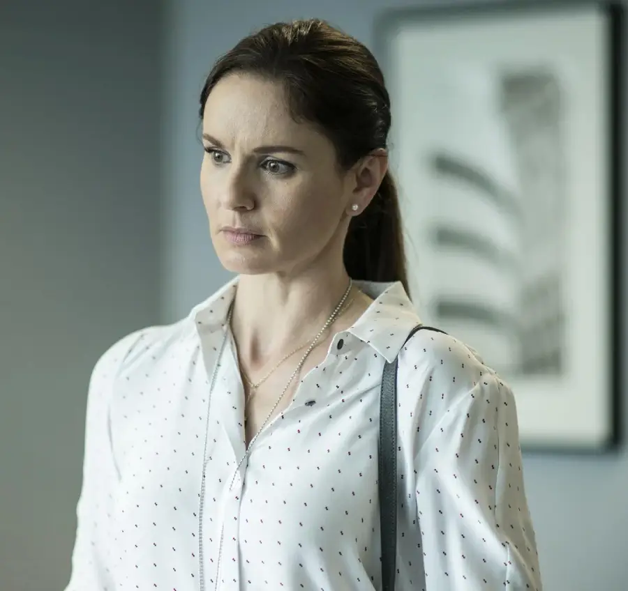 A woman with brown hair in a ponytail, wearing a white blouse with small dots and a necklace, stands indoors looking serious. A blurred black-and-white framed picture is visible in the background.
