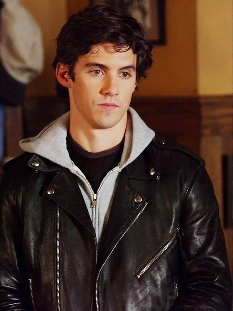 A young man with dark hair wears a black leather jacket over a light gray hoodie, standing indoors against a warm-toned background.