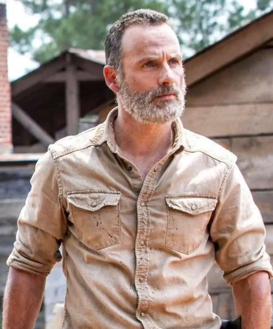 A man with a gray beard and short hair stands outside, wearing a tan button-up shirt with rolled sleeves. Wooden buildings and trees appear in the background.