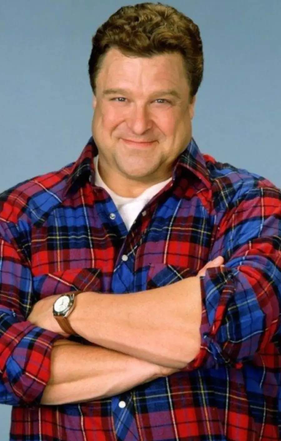 A smiling man with short brown hair wears a red and blue plaid shirt over a white t-shirt. He has his arms folded and stands against a plain blue background.