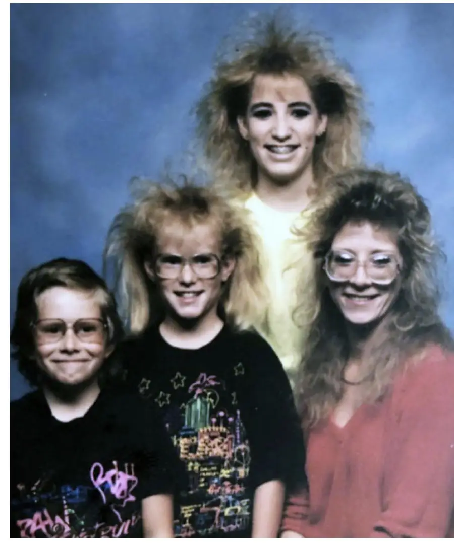 Four people with big, teased 1980s hairstyles and large glasses pose and smile for a studio portrait against a blue background. Three are wearing graphic or colorful tops, and one wears a red sweater.