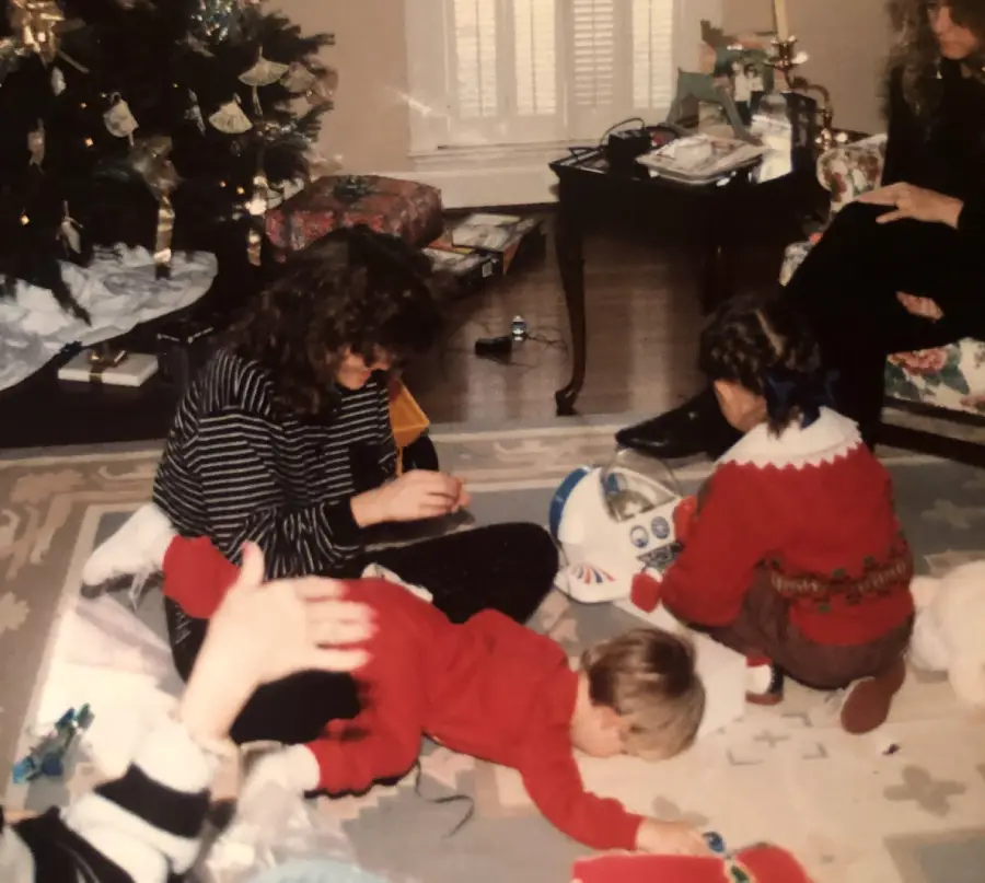 Three children play with toys on a rug near a decorated Christmas tree, while an adult sits on a chair nearby. Wrapping paper and gifts are scattered around the room, suggesting a festive holiday morning.