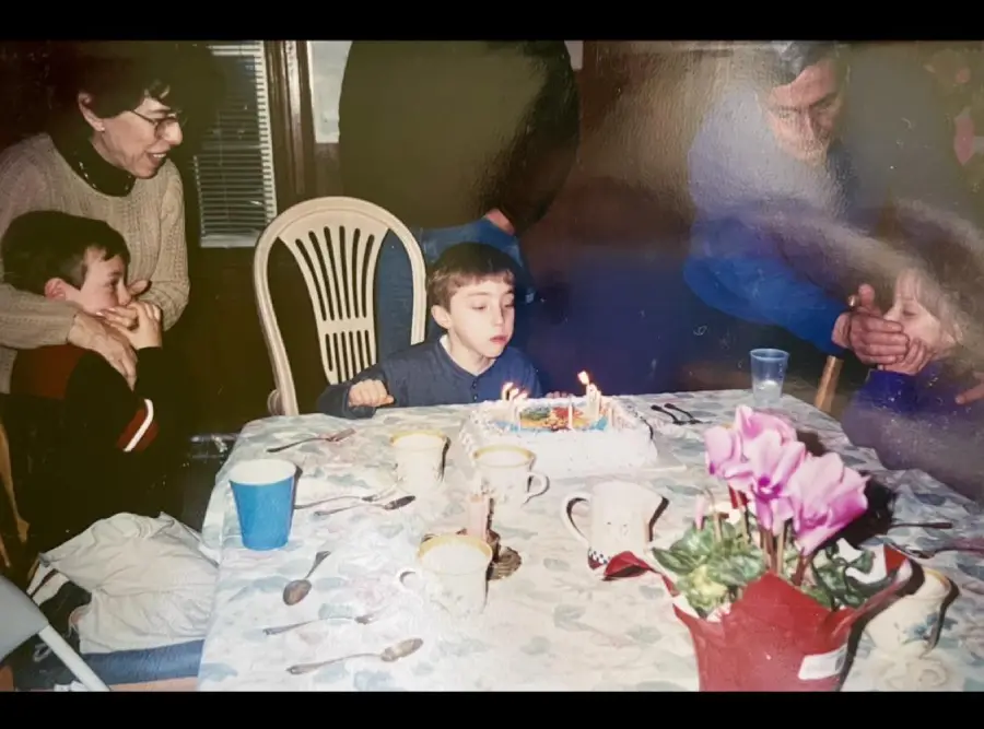 A young boy blows out candles on a birthday cake at a table, surrounded by family members. Cups, mugs, and flowers are on the table, with adults and children watching and smiling.