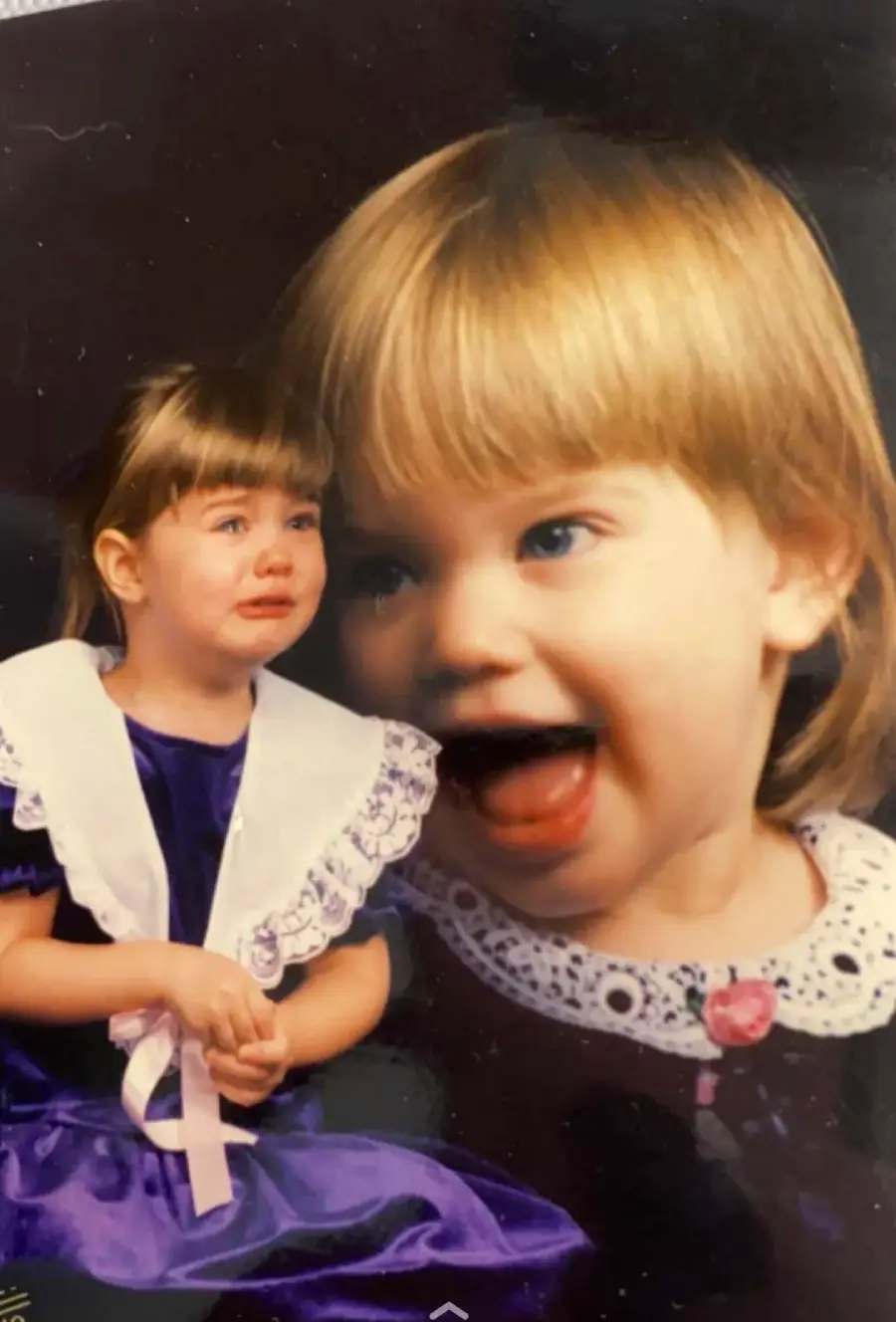 A young girl in a purple dress with a white lace collar sits crying, while a large, faded, smiling portrait of her overlays the background.