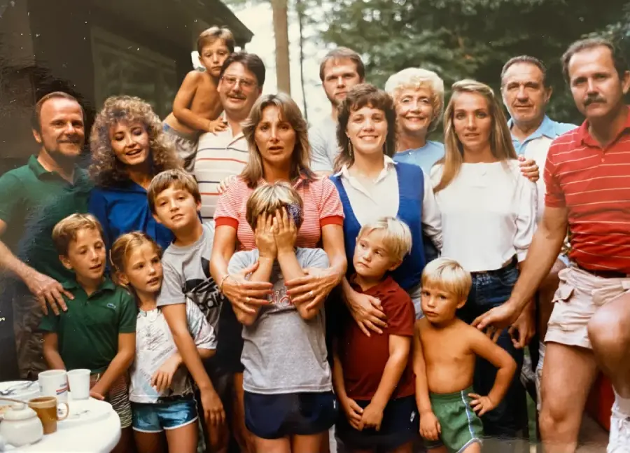 A group of adults and children pose together outdoors, some smiling, some serious. One woman in the center covers a boy’s face with her hands. Trees and patio furniture are visible in the background.