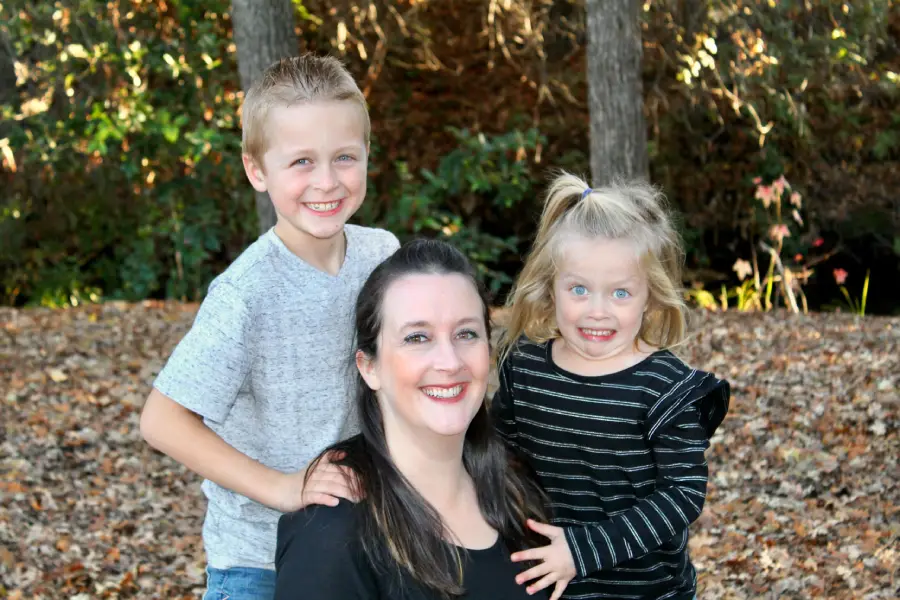 A woman smiles while kneeling outdoors with a young boy standing beside her and a young girl in a black striped shirt hugging her. All three are smiling and surrounded by fallen leaves and trees.