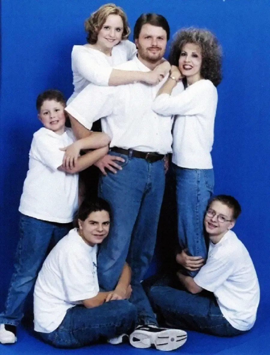 A family of five poses in front of a blue background, all wearing white shirts and blue jeans. The parents stand in the center while their three children, two boys kneeling and one boy standing, hug the parents' legs and waist.