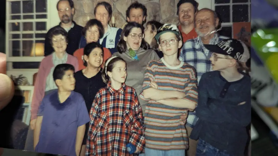 A group of twelve people, both adults and children, pose indoors for a casual photo. Most are smiling or laughing, and some children wear oversized shirts and baseball caps. The background shows a window and a stone wall.