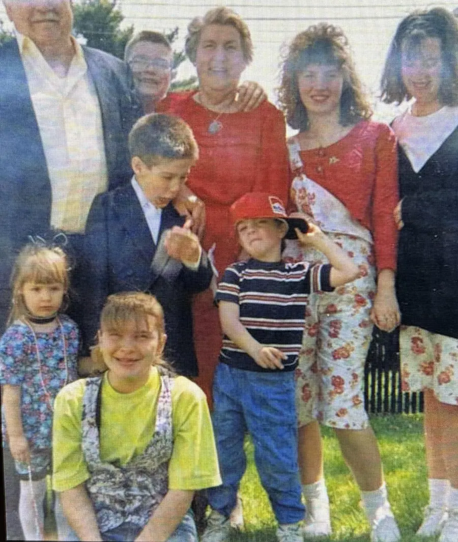 A group of eight people, including adults and children, pose outdoors on a sunny day. Some children smile, while others look away. They are dressed in casual and semi-formal clothes, standing on grass with trees in the background.