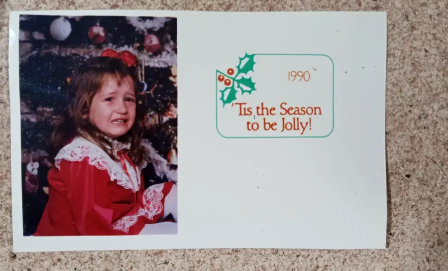 A young girl in a red dress with a white lace collar sits crying in front of a Christmas tree. Next to her, text reads: "1990 'Tis the Season to be Jolly!" with a holly graphic.