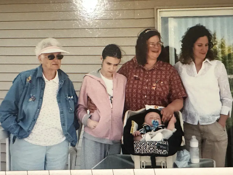 Five people stand in front of a house: an older woman in sunglasses, a teen girl in a pink hoodie, two women in casual clothes, and a baby in a car seat with a bottle beside them. They all face the camera.