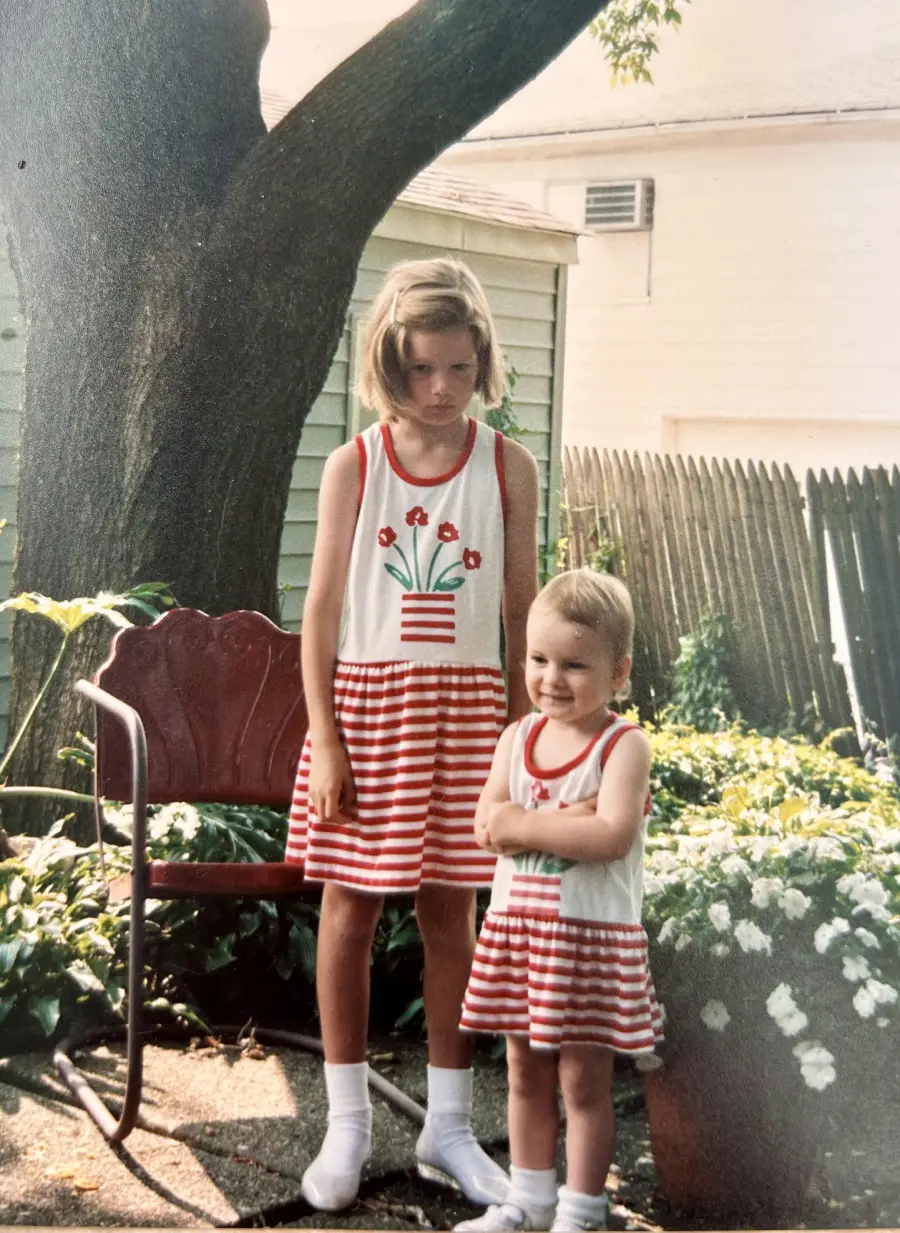 Two young children stand outside in matching white and red striped dresses. The older child looks serious while the younger one smiles. They are near a red chair, flowers, a tree, and a wooden fence in a backyard.