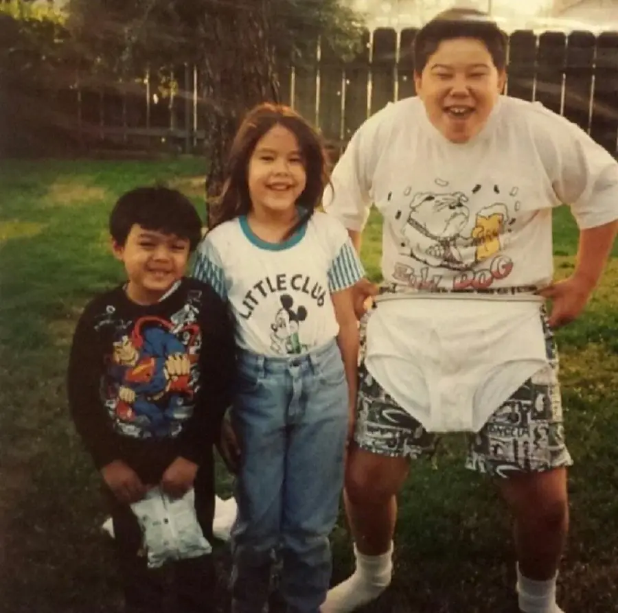 Three children are standing outside on grass, smiling. The child on the right is pulling up and wearing oversized white underwear over their shorts, while the other two stand close, all appearing to enjoy a playful moment.