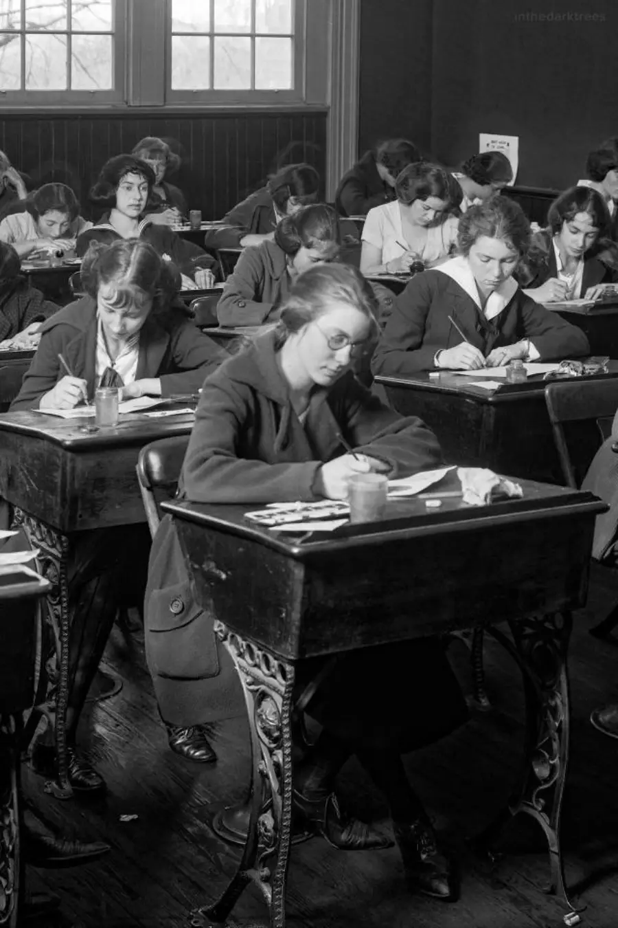 Black and white photo of young women sitting at wooden desks in a classroom, focused on writing or taking a test, with old-fashioned clothing and hairstyles.