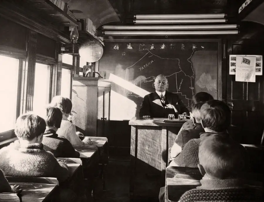 A teacher sits at a desk in front of students in a classroom on a train car, with sunlight streaming in, a globe on a shelf, and a map on the wall behind the teacher.