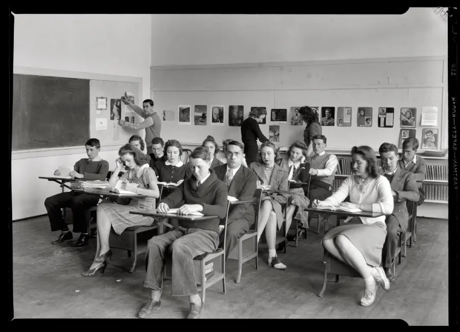 Black-and-white photo of students sitting at desks in a classroom, writing or reading. Two people stand at the back, one drawing on the chalkboard, others looking at art displayed on the wall.