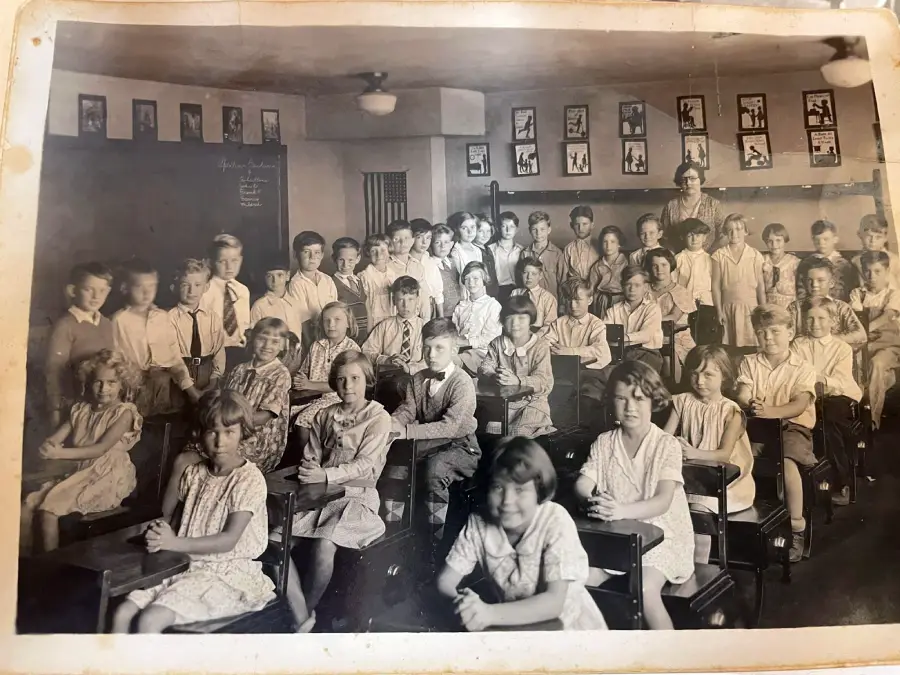 A vintage classroom photo with a large group of children sitting at desks, dressed in old-fashioned clothing, and a teacher standing at the back. The walls display framed pictures. The image is in black and white.