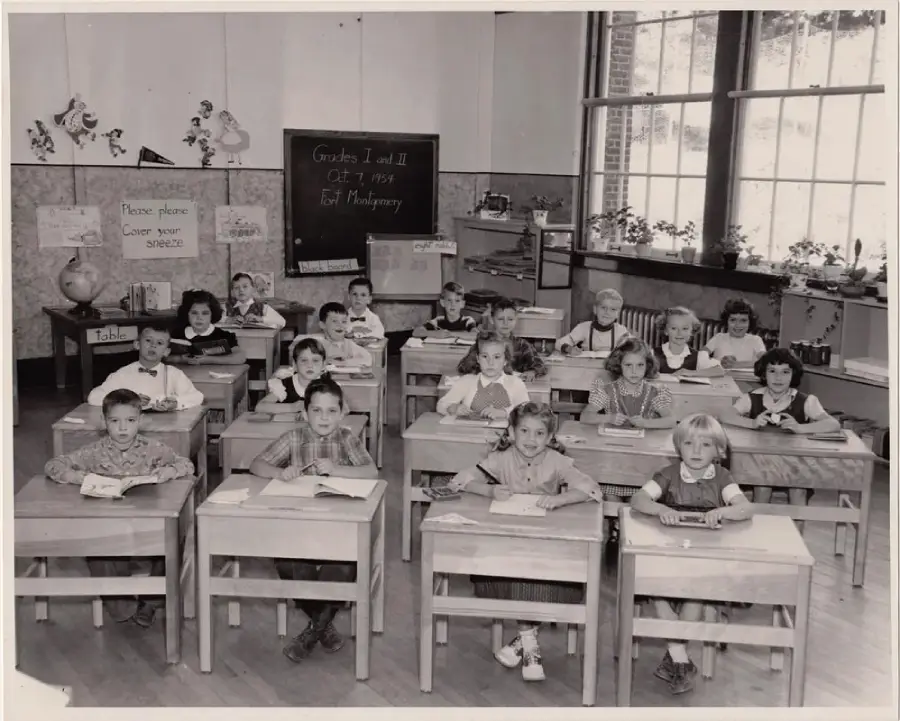 A black-and-white photo of a classroom with children sitting at desks, smiling at the camera. The teacher stands at the back. "Grades I and II Oct 4, 1955" is written on a chalkboard.