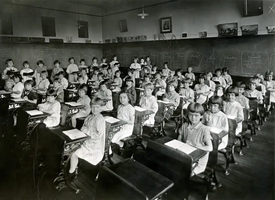 Black and white photo of a classroom filled with young students sitting at desks, facing the camera. The room has chalkboards covered with writing and drawings, and framed pictures on the walls.