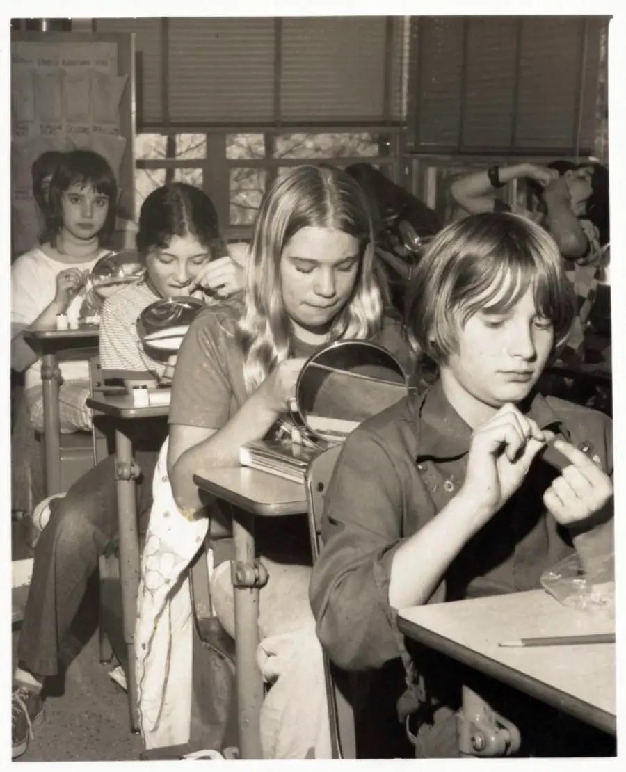 Four students sit in a classroom, each focused on painting their nails or grooming. Desks are arranged in rows, and classroom supplies are visible in the background. The photo is in black and white.