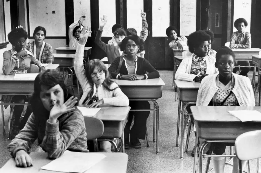Black-and-white photo of students in a classroom sitting at desks. Some have their hands raised. The students appear engaged, and papers are on the desks.
